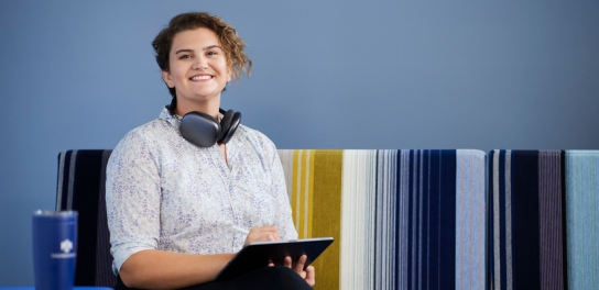 Image of an Undergraduate student wearing headphones and smiling up at the camera while holding a tablet.